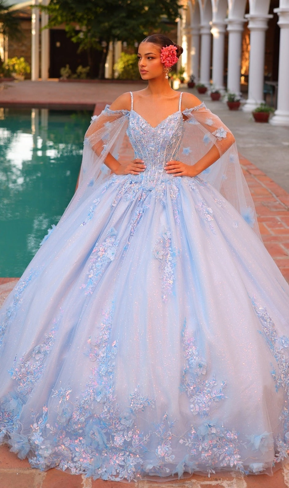 A woman wearing the Quinceanera Dress 54306 By Amarra, featuring a light blue ball gown with a corset bodice and floral embroidery, stands by a pool with flowers in her hair and an ornate building behind her.